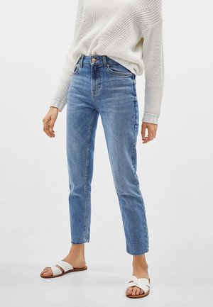 Person wearing light blue high-waist straight-leg jeans, white knitted sweater tucked in, and white flat sandals with crossed straps.