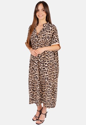 Junge Frau steht und lächelt, trägt ein lockeres Kleid mit Leopardenmuster und schwarze Riemchensandalen vor weißem Hintergrund.