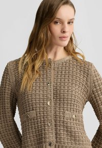 Beige strukturierter Cardigan mit einem Rautenmuster, Rundhalsausschnitt, goldenen Knöpfen und zwei vorderen Taschen. Hergestellt aus einem gewebten Material.
