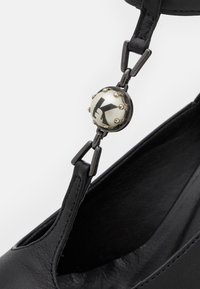 Chaussure en cuir noir avec un bout arrondi. Dispose d'une bride de cheville réglable ornée d'un détail circulaire et de clous, ajoutant texture et intérêt visuel.