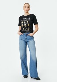 Sort T-shirt med gyldent månekalender grafik, parret med lyseblå brede jeans. Stoffet ser blødt ud, og jeansene har høj talje og bæltestropper.
