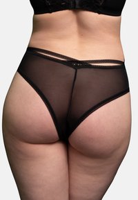 Unterer Rücken und Hüften einer Frau, die durchsichtige schwarze Unterwäsche mit überkreuzten Trägerdetails vor einem einfarbigen Hintergrund trägt.