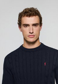 Marinblå stickad tröja med en strukturerad kabelmönster och rund halsringning, med en liten röd logotyp på vänster sida av bröstet.