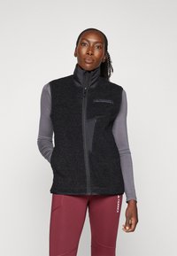 Black fleece vest with high collar, zippered front, and side pockets. Soft texture, contrasting panels, and a logo on the chest.