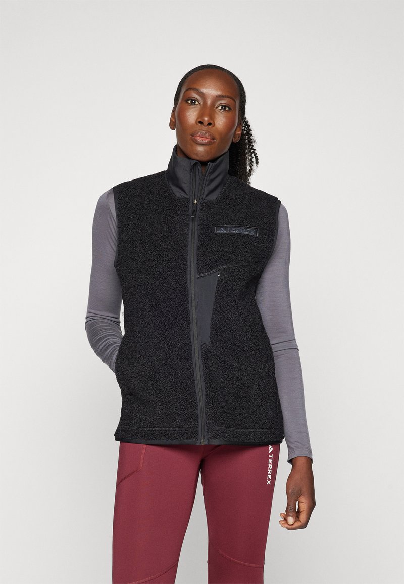 Black fleece vest with high collar, zippered front, and side pockets. Soft texture, contrasting panels, and a logo on the chest.