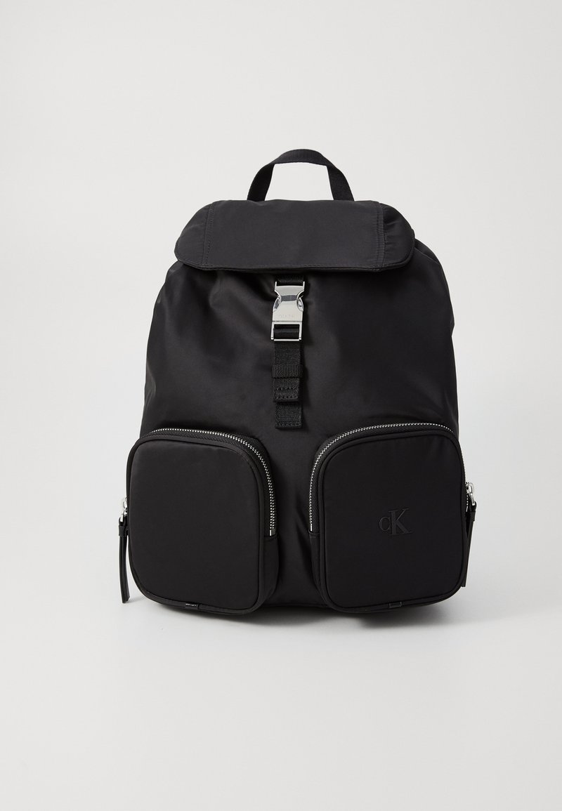 Zaino in nylon nero con apertura a flap, componenti in argento, due tasche frontali con zip e un dettaglio logo testurizzato. Design semplice con spallacci regolabili.