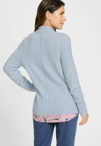 Helles blauer gestrickter Pullover mit hohem Kragen, gerippten Bündchen und geradem Schnitt. Getragen über einem pink- und blau gemusterten Hemd und dunkelblauen Hosen.