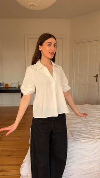 Νεαρή γυναίκα με μακριά σκούρα μαλλιά, φοράει λευκό μπλουζάκι και μαύρο παντελόνι, στέκεται σε ένα φωτεινό υπνοδωμάτιο με ξύλινο πάτωμα και λευκά καλύμματα κρεβατιού.