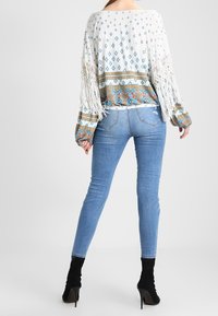 Biała bluzka z długim rękawem z niebieskimi geometrycznymi wzorami, z frędzlami wzdłuż rękawów, zestawiona z jasnoniebieskimi obcisłymi jeansami i czarnymi botkami za kostkę.