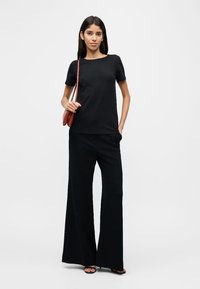 Γυναίκα που φοράει μαύρο κοντομάνικο τοπ και μαύρο παντελόνι φαρδιάς γραμμής, κρατώντας μια μικρή καφέ τσάντα ώμου, στέκεται μπροστά σε λευκό φόντο.