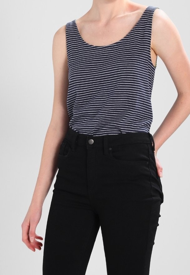 Femme portant un débardeur sans manches rayé bleu marine et blanc rentré dans un pantalon noir taille haute, debout avec une main sur la hanche.
