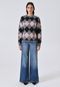 Donna che indossa un maglione argyle grigio, rosa e nero con jeans blu a gamba larga e scarpe nere, in piedi su uno sfondo chiaro e semplice.