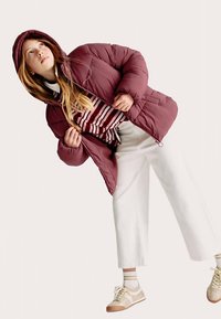 Bordeauxfarvet dunjakke med hætte og lynlåslukning, båret over en stribet skjorte og hvide vidde bukser. Lyse sneakers fuldender looket.