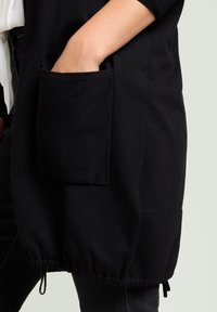 Cardigan long noir à texture tricotée, avec une grande poche latérale et un ourlet froncé. Le tissu est doux, offrant un ajustement confortable.