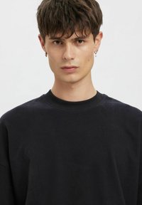 Młody mężczyzna z krótkimi brązowymi włosami, ubrany w czarną bluzę z okrągłym dekoltem i małe kolczyki-koła, zwrócony przodem o neutralnym wyrazie twarzy.