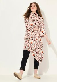 Asymmetrische tuniek met een crèmekleurige basis en een bloemenprint in rood, oranje en paars. Zwarte broek met kantafwerking en lichte sandalen.