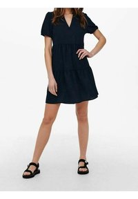 Navyblaues Kurzarmkleid mit V-Ausschnitt, gestuften Design und lockerer Passform, kombiniert mit schwarzen Sandalen mit verstellbaren Riemen.