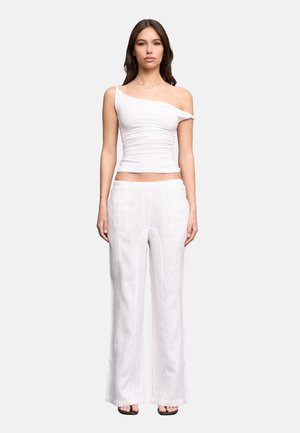 Mujer de pie con top blanco fruncido de un solo hombro y pantalones blancos sueltos con tacones negros abiertos en dedos sobre un fondo liso.