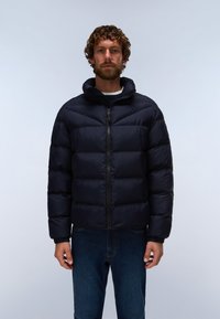 Man met krullend haar en baard draagt een zwarte donsjas en blauwe jeans, staand tegen een effen lichte achtergrond.