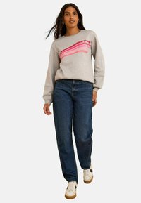 Sudadera gris con diseño de corazón a rayas rosas, combinada con unos vaqueros azul oscuro y zapatillas blancas con detalles marrones. Ajuste casual, de mangas largas.