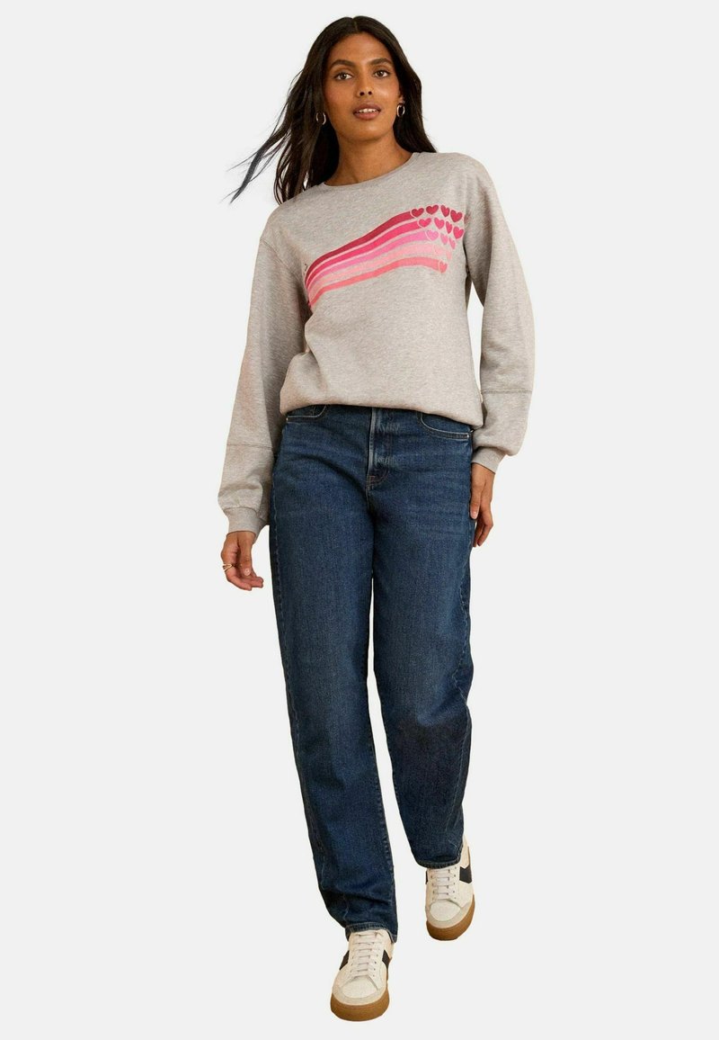Sudadera gris con diseño de corazón a rayas rosas, combinada con unos vaqueros azul oscuro y zapatillas blancas con detalles marrones. Ajuste casual, de mangas largas.