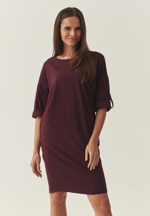 Kobieta o długich brązowych włosach, ubrana w luźną, do kolan, bordową sukienkę z podwiniętymi rękawami, stojąca na jednolitym tle.