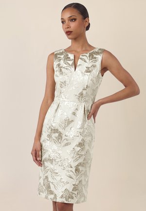 Frau trägt ärmelloses, knielanges cremefarbenes Kleid mit metallischem goldfarbenem Blumenmuster, eine Hand in der Hüfte, schaut seitlich vor einfachem Hintergrund.