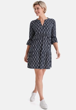 Frau, die ein marineblaues und weißes geometrisch gemustertes Kleid mit dreiviertellangen Rüschenärmeln und weißen Turnschuhen trägt und geht.