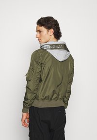 Olivgrüne Bomberjacke mit grauem Kapuzen. Verfügt über elastische Bündchen und Saum, Reißverschlusstasche am Ärmel und Logodruck auf der Rückseite der Kapuze.