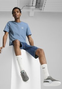Šviesiai mėlynas medvilninis marškinėliai su PUMA logotipu, džinsiniai šortai, pilki zomšiniai kedai ir balti kanalų rašto kojinės su juodais logotipo akcentais.