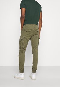 Olivengrønne cargo-bukser med elastiske manchetter, flere lommer og en afslappet pasform, parret med hvide sneakers og en mørkegrøn t-shirt.