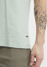 Camiseta de manga corta verde claro con dobladillo redondo, hecha de una tela suave, que presenta una pequeña etiqueta blanca con un logo negro en el lado inferior.