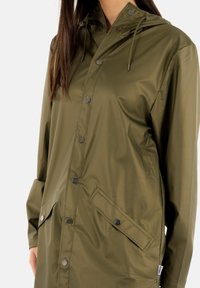 Chaqueta impermeable de color verde oliva, con capucha, botones a presión, cordones ajustables y un bolsillo frontal con cierre a presión. Textura de tela suave.