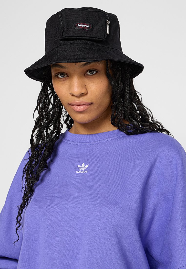 Un chapeau seau noir avec une poche avant et une fermeture éclair, fabriqué en tissu. Porté avec un sweat-shirt violet avec un petit logo blanc.