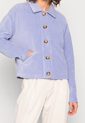 Chaqueta de pana lila claro con grandes botones y bolsillos frontales, usada sobre pantalones blancos de corte holgado.