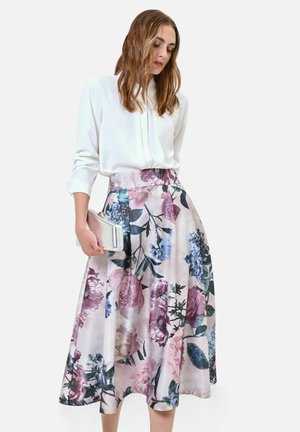 Femme en chemisier blanc à manches longues et jupe midi fleurie taille haute tenant une pochette blanche sur un fond uni.