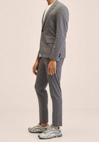 Man in een grijs pak met tapse broek, een wit T-shirt en lichtgrijze sport sneakers, staande tegen een effen beige achtergrond.