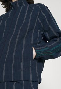 Marinblå jacka med vertikala ränder, med hög krage, dragkedjestängning, texturerad tyg och gröna detaljer på ärmarna.