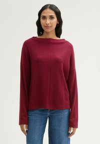 Burgunderfarbener Strickpullover mit einem Bootsausschnitt, überschnittenen Schultern und langen Ärmeln. Kombiniert mit blauen Jeans aus Denim. Strukturiertes Material.