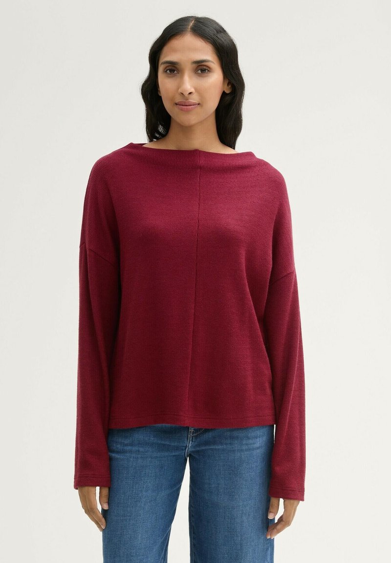 Burgunderfarbener Strickpullover mit einem Bootsausschnitt, überschnittenen Schultern und langen Ärmeln. Kombiniert mit blauen Jeans aus Denim. Strukturiertes Material.