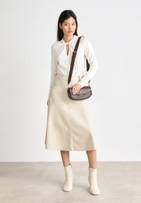 Beige skjørt med knelange detaljer, kremfarget langermet topp med nøkkelhull, og en brun crossover-veske. Jevn tekstur og minimalistisk design.