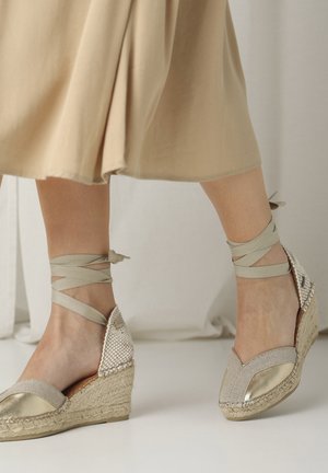 Alpargatas de cuña con parte superior de tejido trenzado, acentos dorados y tela color beige. Las correas del tobillo están atadas alrededor del tobillo para un ajuste seguro.