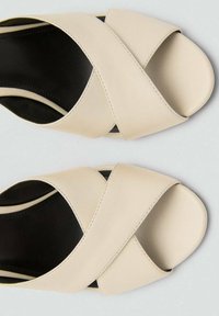 Sandalias beige de tira cruzada, fabricadas en cuero suave con una suela ligeramente texturizada. Diseño destalonado con forro negro interior y detalles de costura estrecha.