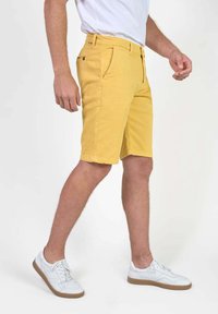 Shorts jaunes en coton, dotés d'une fermeture éclair à l'avant, de poches latérales et d'une coupe droite. Portés avec des baskets blanches.