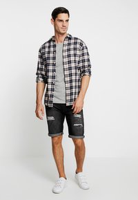 Camicia in flanella a quadri nei colori blu navy, crema e rosso; t-shirt grigia; pantaloni corti in denim nero strappati; sneakers bianche. Outfit casual con le maniche arrotolate.