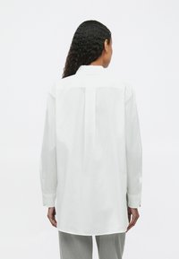 Cămașă albă cu mâneci lungi și manșete cu butoane, detaliu cu pliu la spate și guler clasic. Material din bumbac neted, croială lejeră.