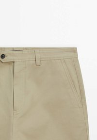 Beige pants with front pockets, belt loops, and a single black button closure on plain white background.
