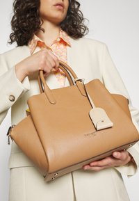 Kobieta w kremowej marynarce trzyma torebkę z brązowej skóry od Kate Spade z wyraźnymi uchwytami i kremową zawieszką.
