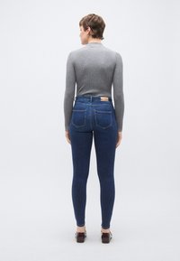 Στενά τζιν με ψηλή μέση σε σκούρο μπλε χρώμα με πίσω τσέπες, συνδυασμένα με γκρι ριμπ μακρυμάνικη μπλούζα. Ορατά επίπεδα παπούτσια.