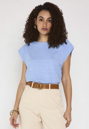 Donna con capelli ricci che indossa un top azzurro senza maniche, pantaloni beige con cintura marrone e orecchini a cerchio e braccialetti dorati.